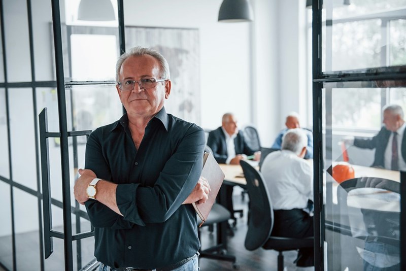 Portrait of a senior man that stands in front of a team of elderly businessman architects that are having a meeting in the office