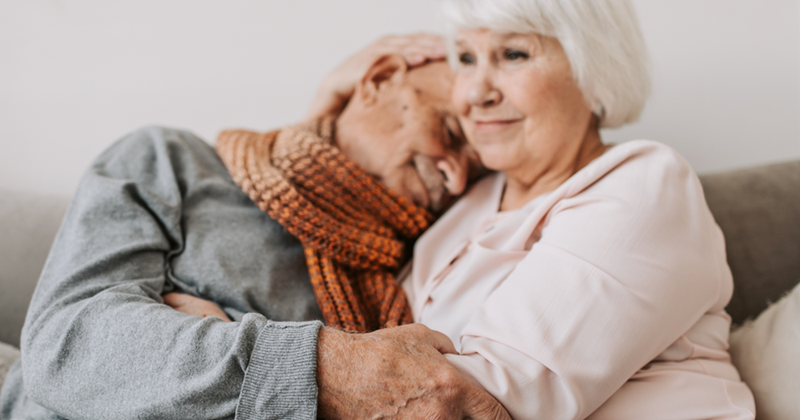 Elderly couple sitting on a couch embracing, with the man resting his head on the woman’s shoulder in a tender moment.