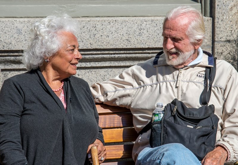 Elderly couple sitting together on a park bench, smiling and chatting while enjoying a sunny day outdoors.