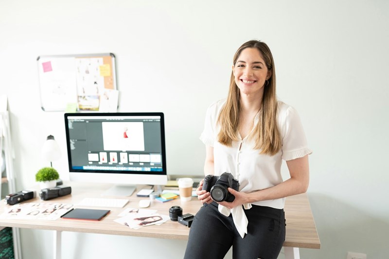 Smiling woman photographer holding a DSLR camera while sitting on a desk in a bright home office with photo editing software open on a computer.