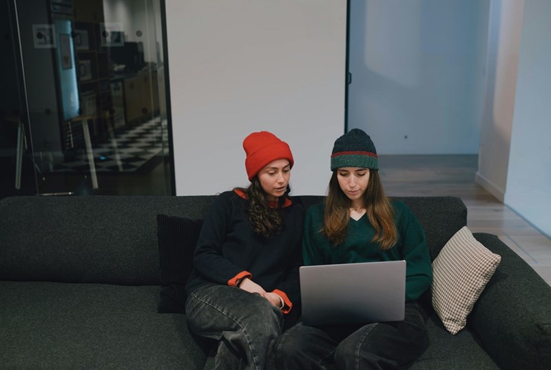 Two women wearing beanies sit on a couch complaining about their coworkers blunt emails