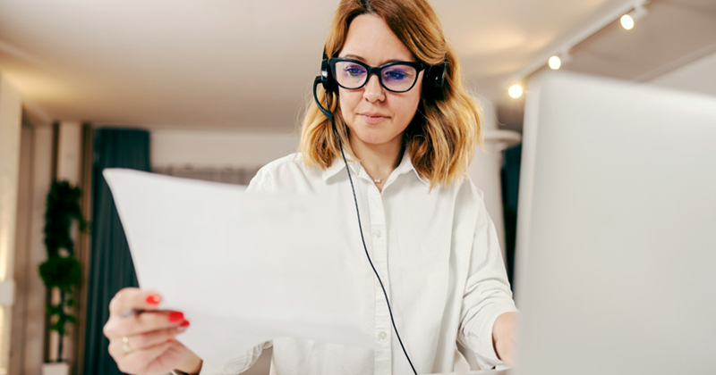A woman wearing glasses and a headset holds up some papers while sitting at her laptop