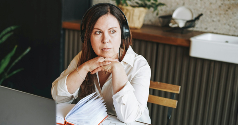 A woman wearing headphones with an open notebook and laptop in front of her looks away thoughtfully