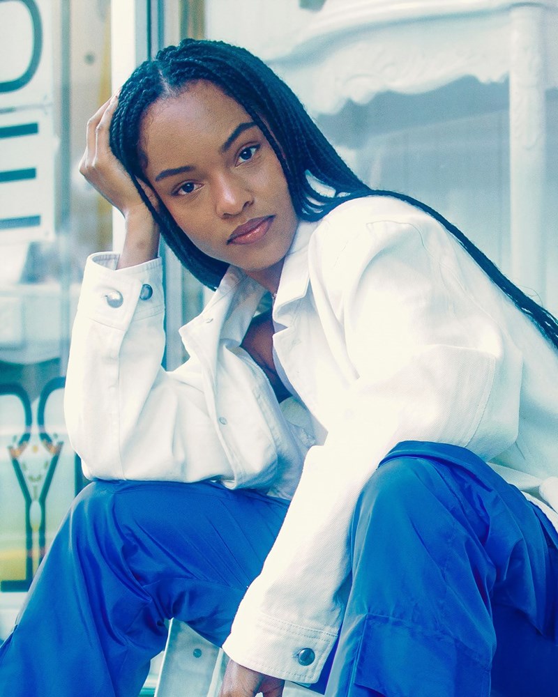 Thoughtful young woman with braided hair sitting by a window, resting her head on her hand while gazing off to the side.