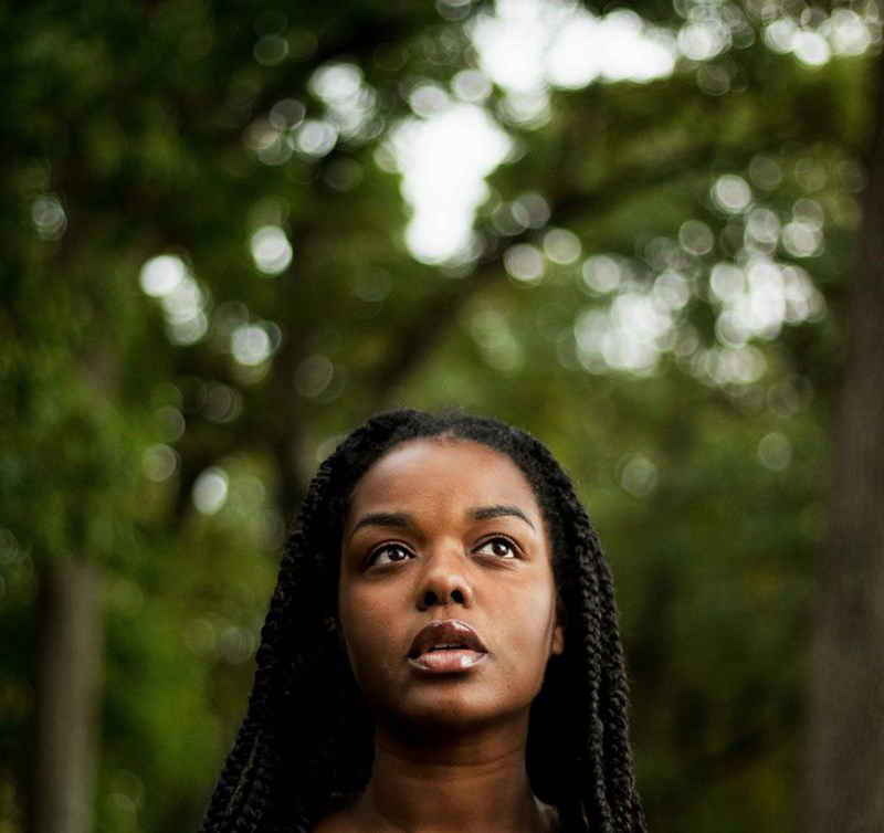 Young woman with braided hair looking upward outdoors, standing in front of blurred green trees in a park setting.