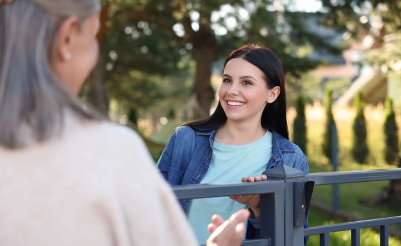 Smiling woman talking with an older neighbor over a backyard fence on a sunny day in a residential neighborhood.