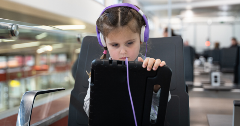 Little girl is playing on an iPad tablet with headphones on at the airport. 