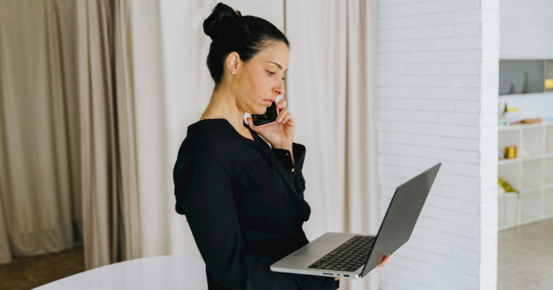 A standing woman dressed in black takes a call on her cell phone while holding her laptop