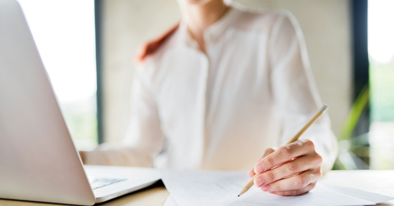 A woman in a white shirt writes on a piece of paper while using a laptop