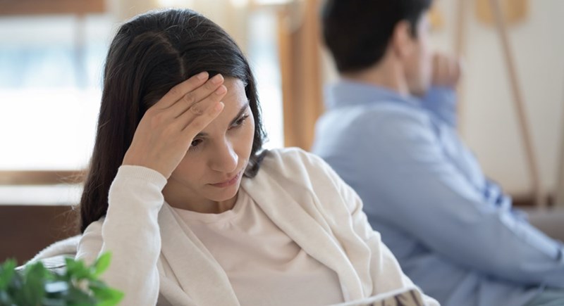 Stressed woman sitting on a couch with her hand on her forehead while a man sits in the background facing away after an argument.