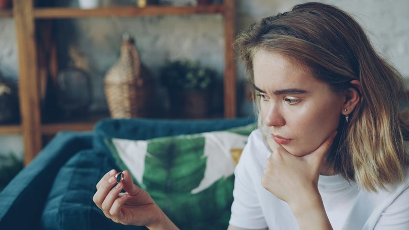 Thoughtful young woman sitting on a couch while holding and examining a ring, looking concerned or conflicted.