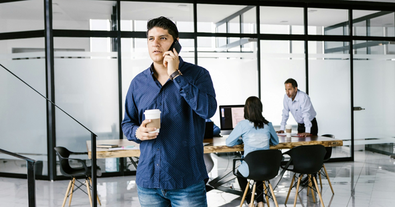 A male office worker holds a coffee and talks on his cell phone in an open plan workspace