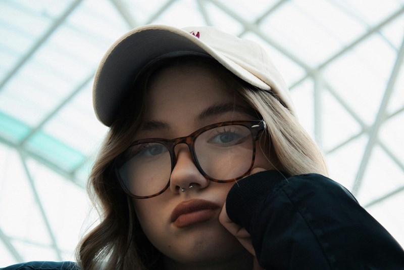 Close-up of a young woman wearing glasses and a baseball cap, resting her chin on her hand with a bored or pensive expression.