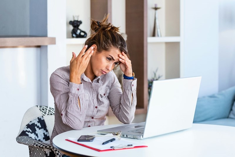 Stressed woman sitting at a table with a laptop, holding her temples while looking overwhelmed by work or bills at home.