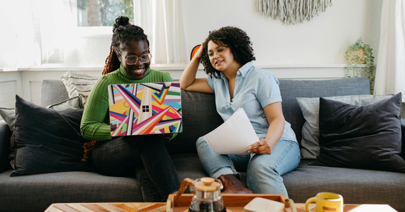 Two women sit on a couch, one holding papers and the other working on a laptop