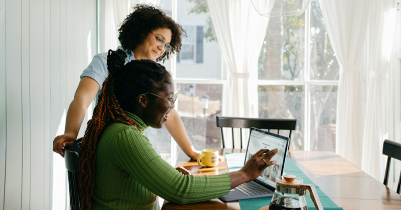 A woman stands over another woman, who is pointing to something on her laptop on a kitchen table