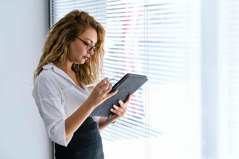Young employee holds a tablet while taking notes next to a window