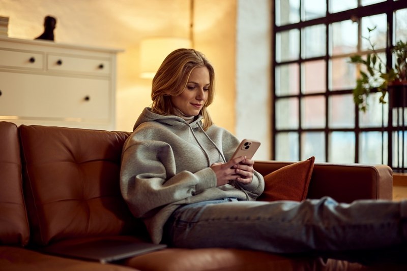 Relaxed woman sitting on a couch in a warm living room, smiling slightly while using her smartphone.