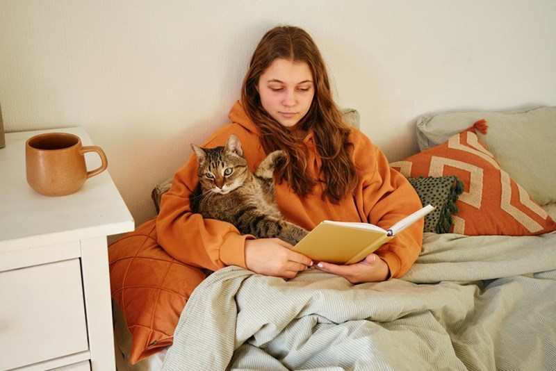 A woman sits in bed reading a book while her cat rests comfortably on her lap in a cozy room.