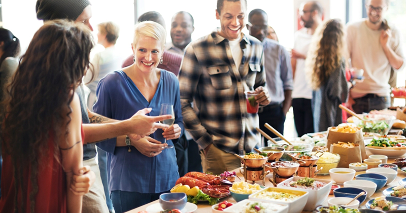 A group of people, some holding beverages, gather around a large buffet table