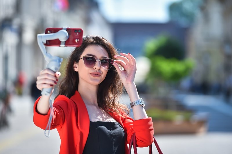Stylish woman wearing sunglasses and a red blazer taking a selfie with a smartphone on a handheld gimbal while standing on a sunny city street.