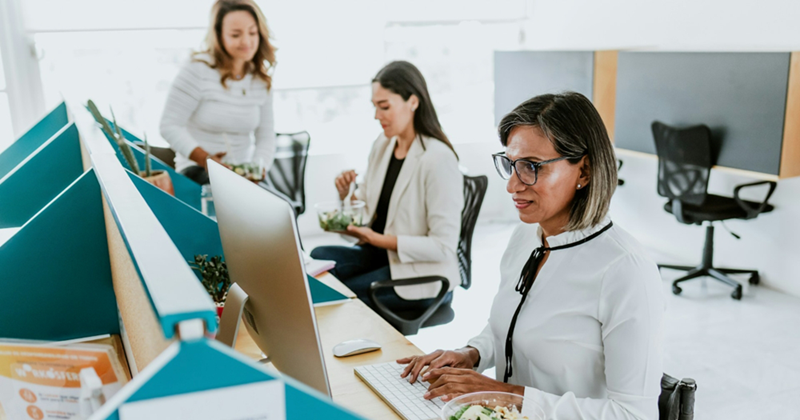 An older woman works at a computer while two colleagues eat salad at their desks behind her