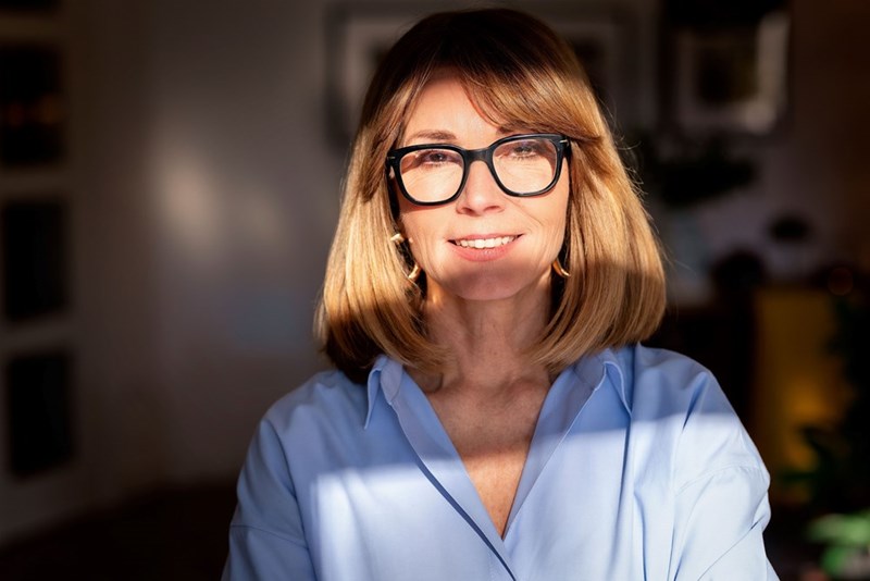 Smiling middle-aged woman wearing glasses and a blue shirt, warmly lit by sunlight indoors.