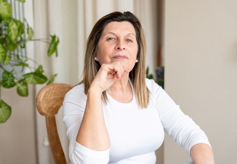 Confident middle-aged woman sitting at a table with her hand under her chin in a bright home interior with plants.