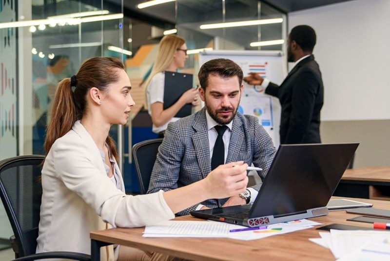 Two coworkers discussing data on a laptop during a meeting in a modern office while colleagues review charts in the background.