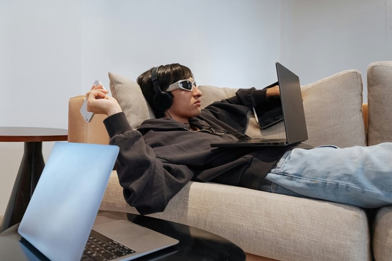 A man in sunglasses lies on the couch while working on his laptop