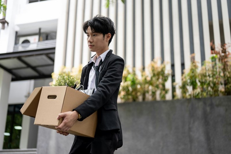 Ashamed worker leaves workplace he's been fired from while holding box of his possessions.