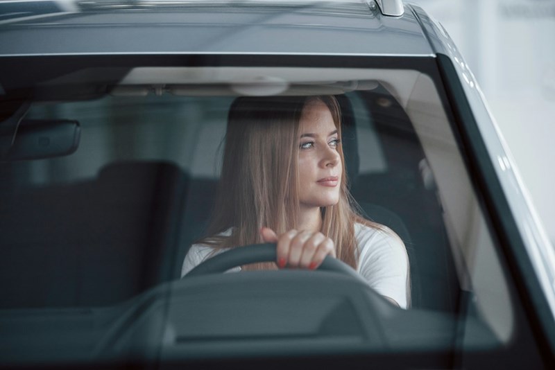 A woman sits behind the wheel, looking off to the side as she holds the steering wheel, appearing calm and focused before driving.