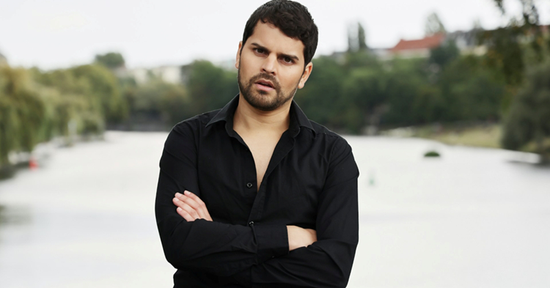 A bearded man stands on a bridge outside with his arms crossed