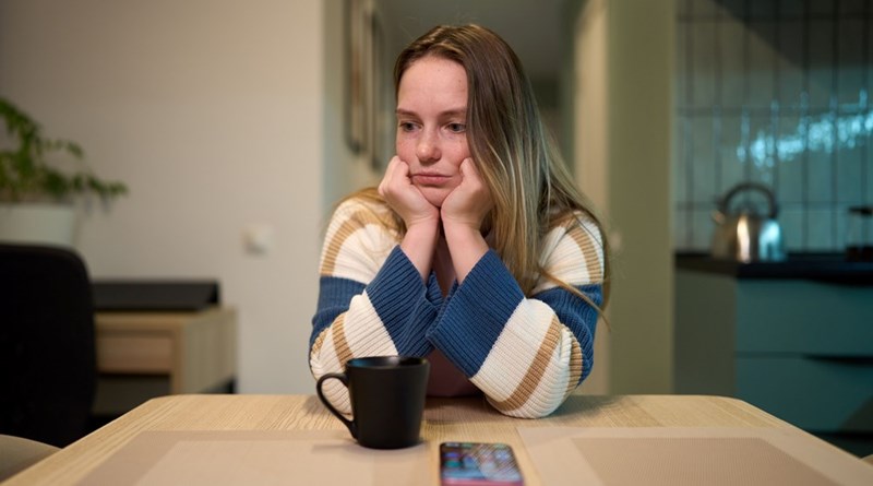Young woman resting her chin on her hands at a table, looking wooried with a mug and smartphone in front of her.