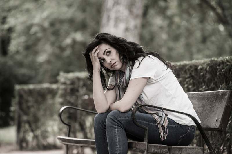 A woman sitting alone on a park bench, leaning forward, her head held in her hand. She has an exasperated expression. The picture is diluted and the atmosphere is grey.
