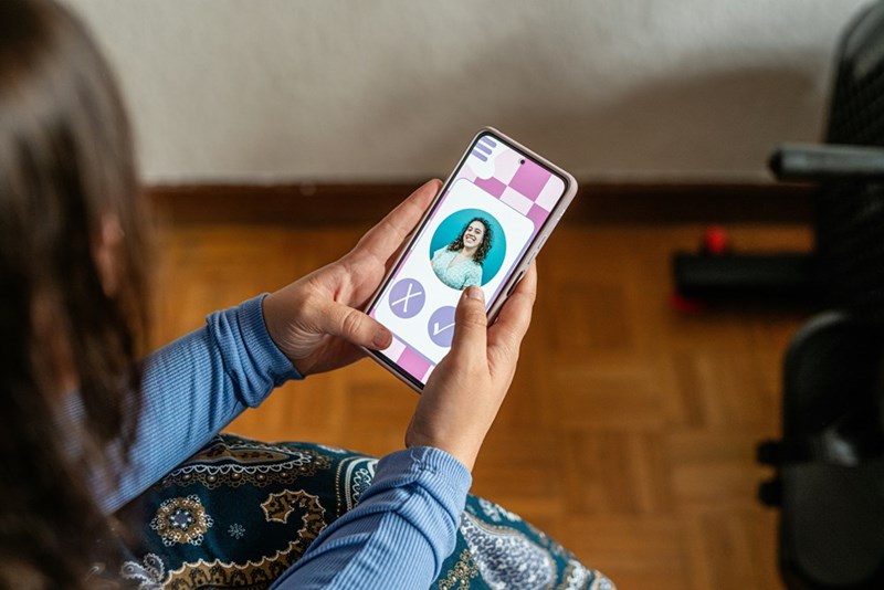 A person holding a phone and swiping through a dating app profile while sitting indoors.