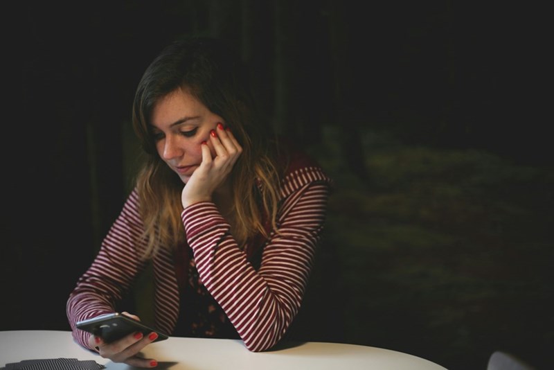 Young woman sitting at a table in low light, resting her head on her hand while looking at her phone.