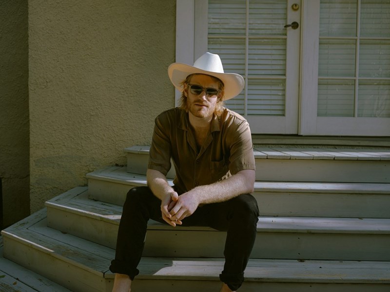 Country man sitting on his home's front porch wearing sunglasses and a cowboy hat looking concerned.