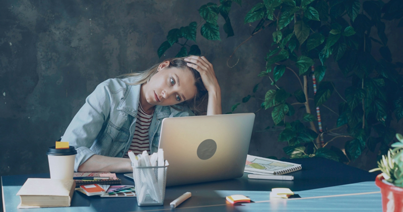 A female employee surrounded by office stationery runs her fingers through her hair as she looks at her laptop screen