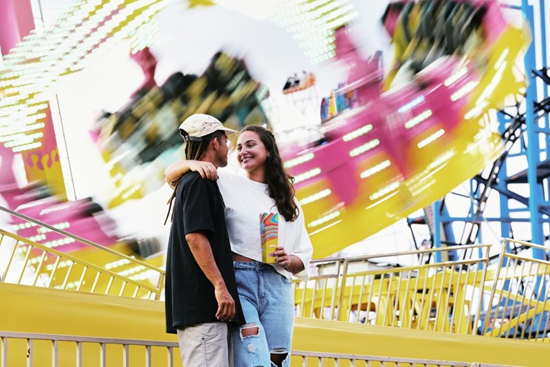 Woman puts arm around her boyfriend as amusement park ride swings behind them.