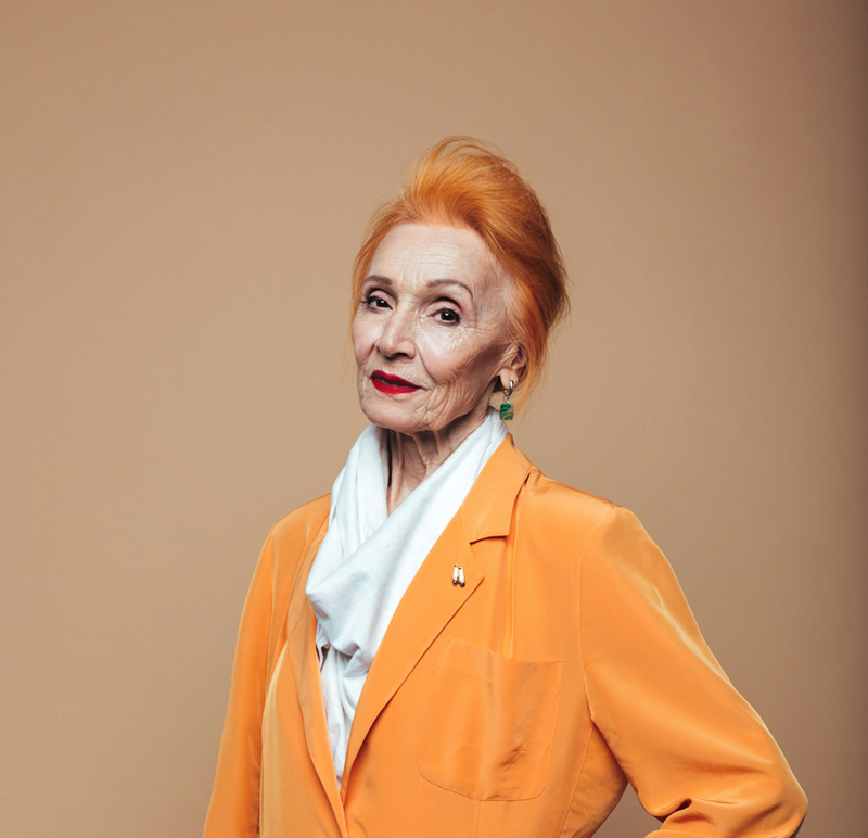 An finely dressed older woman in an orange shirt gazes intensely back with a piercing glare