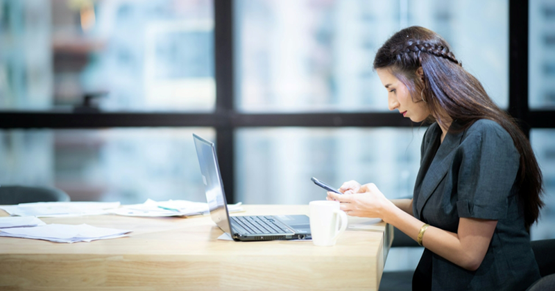 A female employee texts on her phone while sitting in front of her laptop in the office
