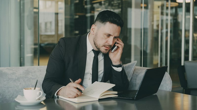 A man in a suit talks on the phone while writing in a notebook and looking at his computer