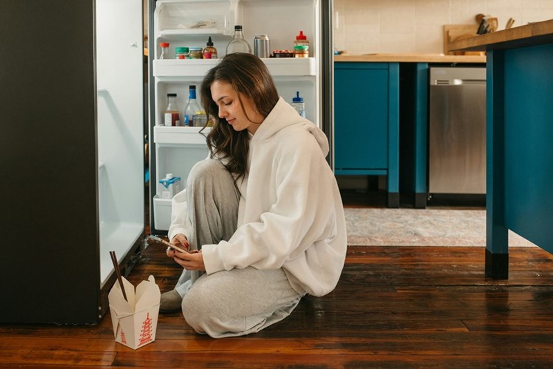 Woman sitting on the kitchen floor by an open fridge, looking at her phone with a takeout box beside her.