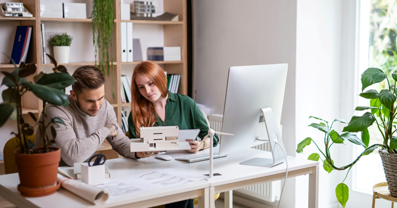 A man and a woman look at an architectural model that the man is holding up while sitting at a desk in a personal office