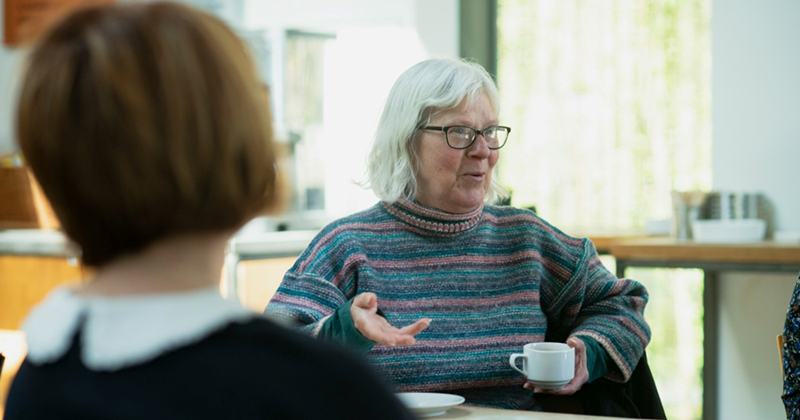 An older woman sitting at a table and holding a coffee cup tells a story to others at the table