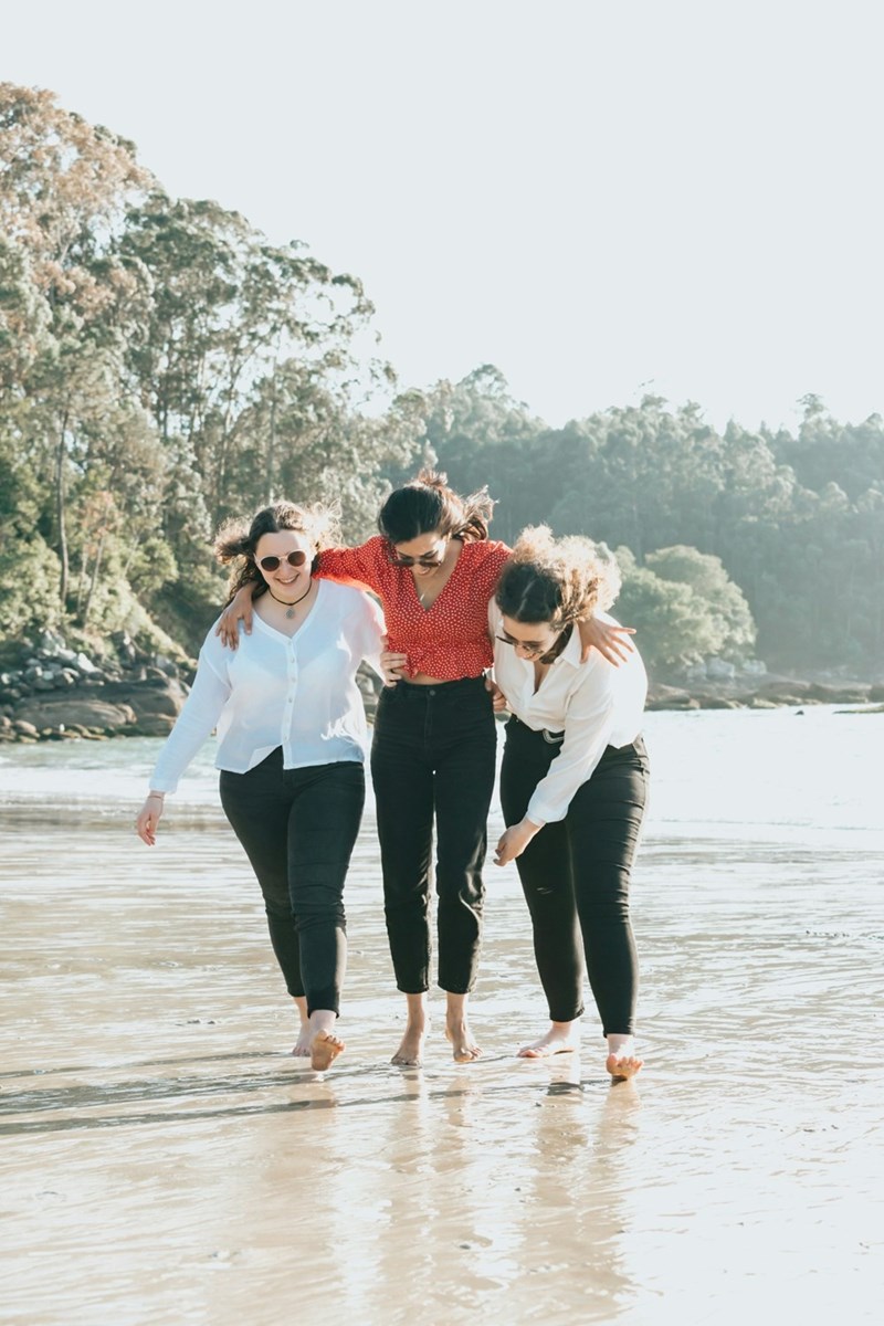 Three female friends put their arms around one another as they walk barefoot across a beach