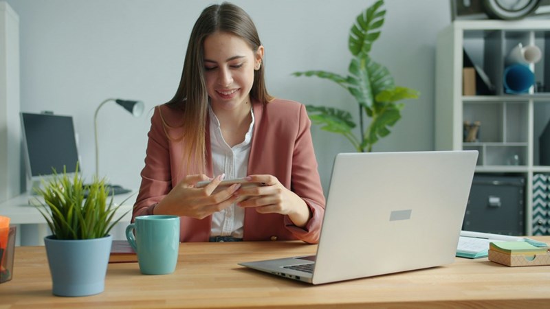 A woman works at her desk checking her phone and her laptop