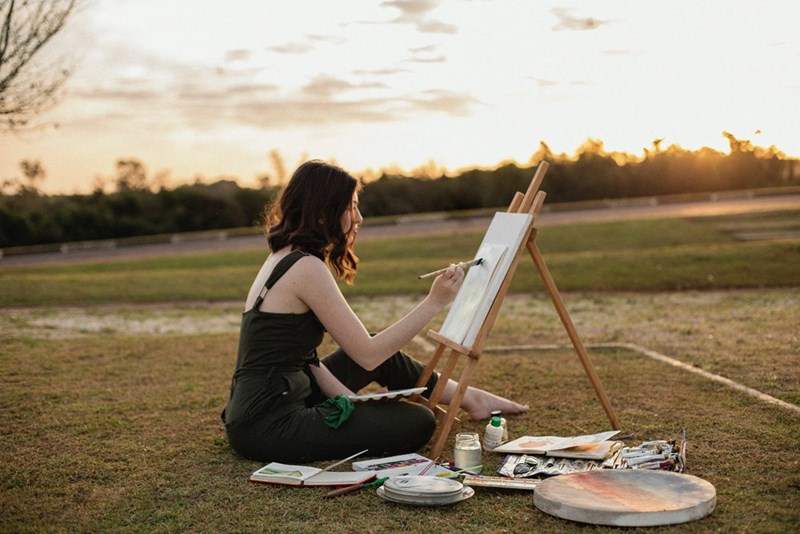 Young woman in black overalls paints a picture for an art competition while relaxing on the ground outdoors.