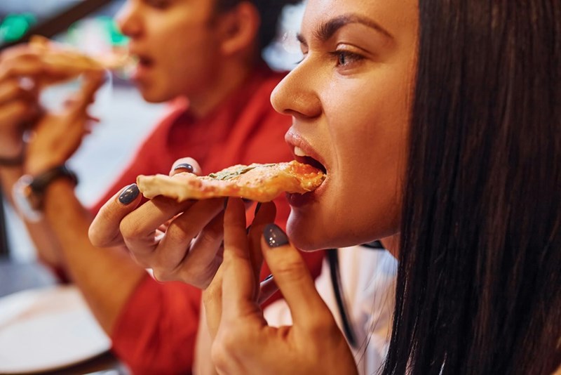 A woman eats a slice of pizza at a restaurant with a friend next to her